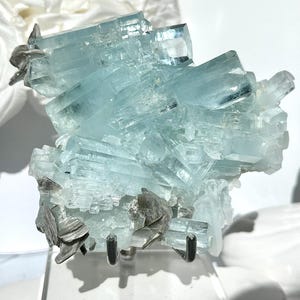 May include: A cluster of aquamarine crystals, showcasing a pale blue hue with crystalline structures. The mineral specimen is mounted on a clear stand, with dark gray accents. The background features white roses and a white sculpture.