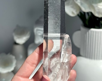 May include: A large, clear quartz crystal point with a double termination. The crystal is held in a hand and has a natural, raw appearance.