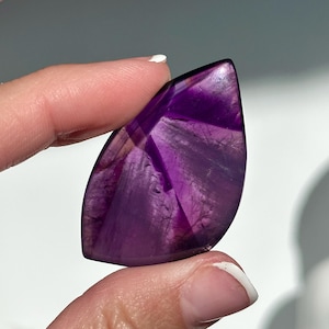 May include: A polished, teardrop-shaped amethyst gemstone. The purple stone has a translucent quality, with visible internal patterns and variations in color intensity. The gemstone is held between two fingers.