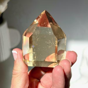 Puede incluir: Una punta de cristal facetada de color amarillo dorado con una punta marrón rojizo. El cristal se sostiene en una mano, mostrando su forma geométrica y su calidad translúcida. La superficie del cristal refleja la luz, creando un efecto brillante.