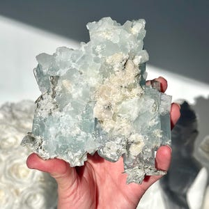 May include: A light blue aquamarine crystal cluster with a rough, textured surface. The crystal has areas of white and tan coloration, and is held in a hand. The crystal is a natural mineral specimen.
