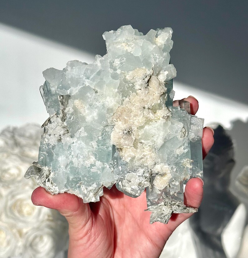 May include: A light blue aquamarine crystal cluster with a rough, textured surface. The crystal has areas of white and tan coloration, and is held in a hand. The crystal is a natural mineral specimen.