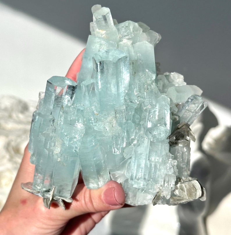 May include: A cluster of aquamarine crystals, showcasing a pale blue hue. The crystals are held in a hand, revealing their natural, irregular formation. The light reflects off the facets, creating a sparkling effect. The overall appearance is of a raw, natural mineral specimen.