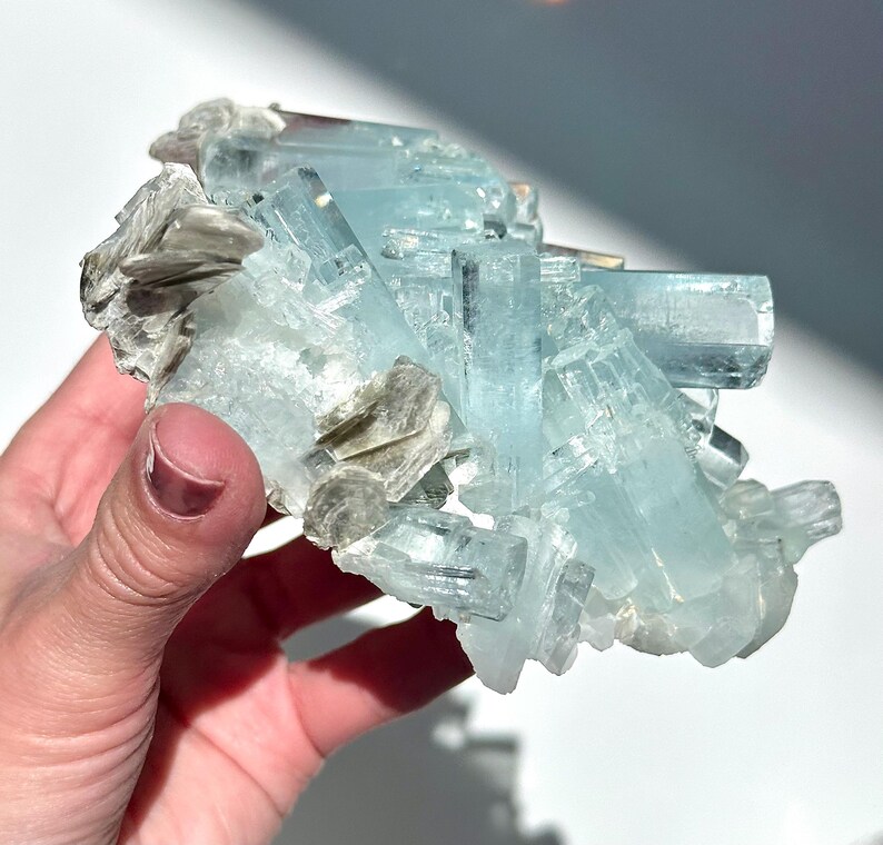 May include: A cluster of aquamarine crystals, showcasing a pale blue hue, with a matrix of silvery-gray mica. The crystals are translucent and have a natural, raw appearance. The mineral specimen is held in a hand.