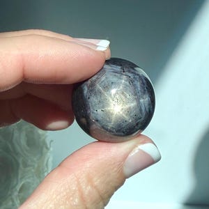 May include: A polished, spherical gemstone with a star-like pattern. The stone is primarily dark gray with lighter gray and gold-colored inclusions. The gemstone is held between two fingers, showcasing its reflective surface and unique design.