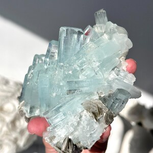 May include: A cluster of aquamarine crystals, showcasing a pale blue hue. The crystals are arranged in a layered formation, with some appearing as elongated prisms. The mineral specimen is held in a hand, with a neutral background.