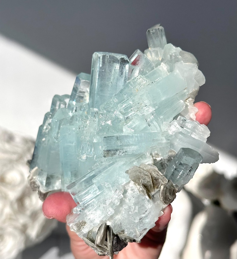 May include: A cluster of aquamarine crystals, showcasing a pale blue hue. The crystals are arranged in a layered formation, with some appearing as elongated prisms. The mineral specimen is held in a hand, with a neutral background.