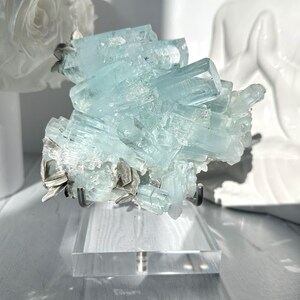 May include: A cluster of pale blue aquamarine crystals on a clear acrylic stand. The crystals are multifaceted and arranged in a layered formation. The stand has a metal support. A white rose and a white statue are in the background.