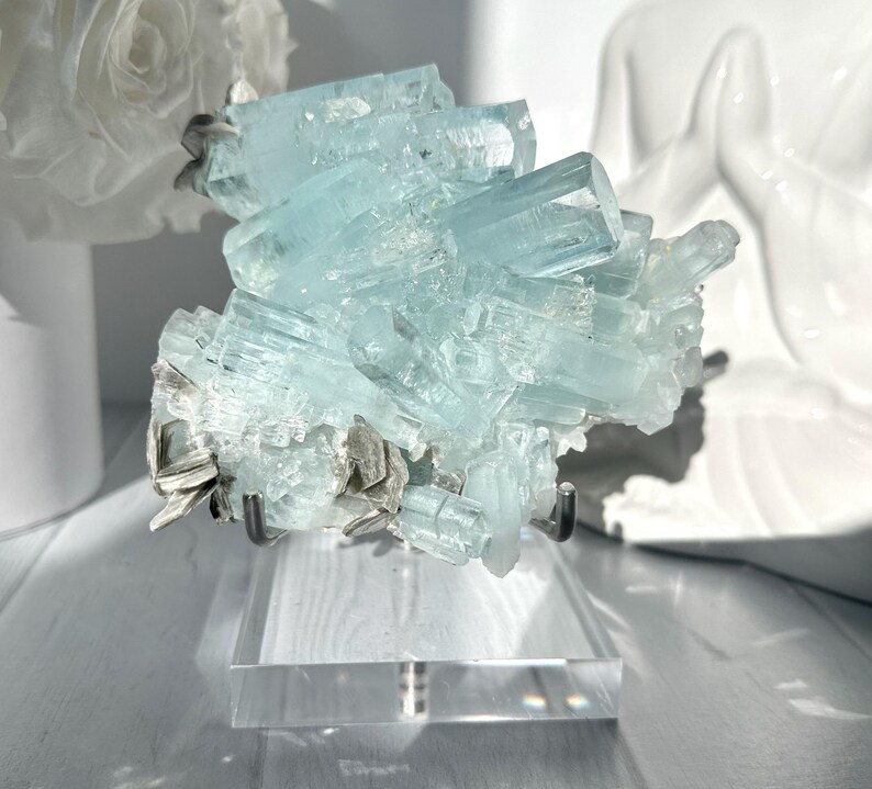 May include: A cluster of pale blue aquamarine crystals on a clear acrylic stand. The crystals are multifaceted and arranged in a layered formation. The stand has a metal support. A white rose and a white statue are in the background.