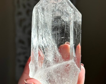 May include: A clear quartz crystal point with a rough, natural texture. The crystal is held in a hand, showcasing its size and clarity.