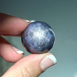 May include: A round, polished gemstone with a star-like pattern. The gem is held between two fingers, showcasing its spherical shape and the play of light across its surface. The stone's color is a mix of blue and silver.