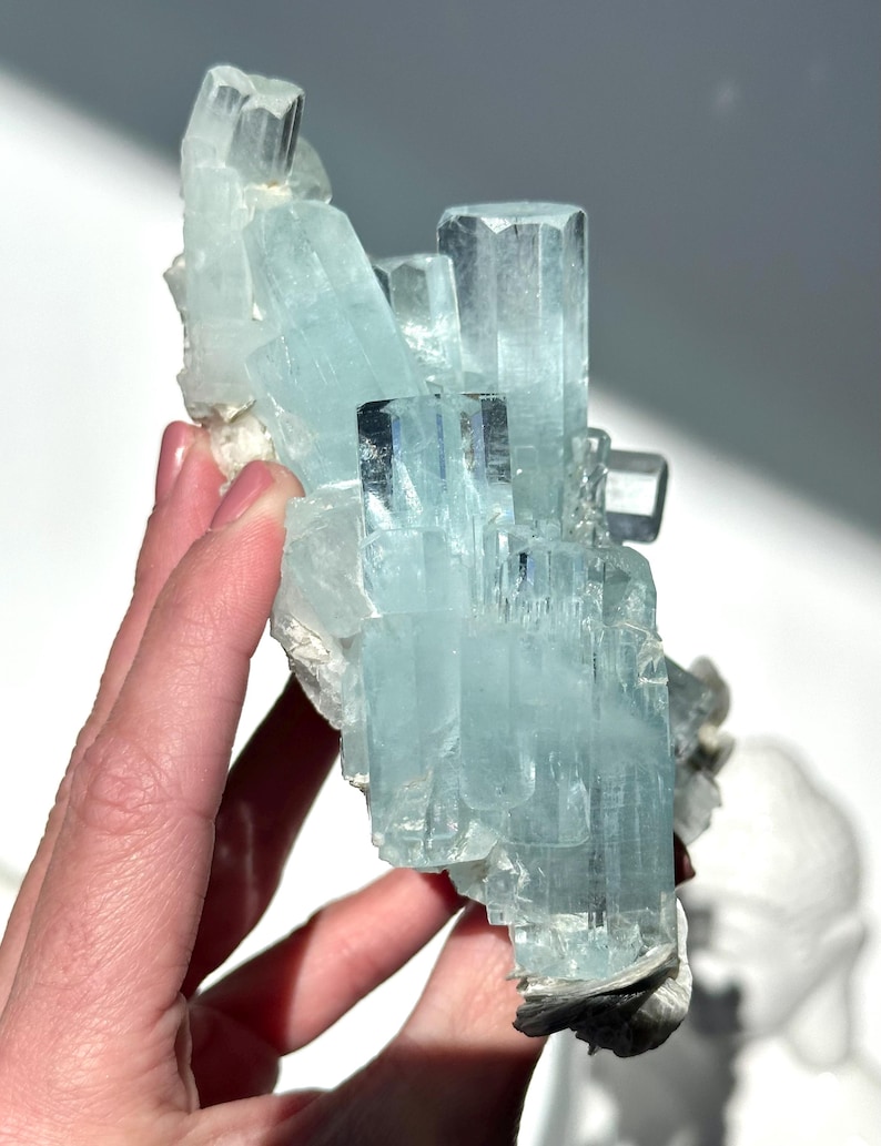 May include: A cluster of aquamarine crystals, showcasing a pale blue hue with crystalline structures. The mineral formation is held in a hand, revealing its natural, raw form. The crystals have a translucent quality, catching the light.