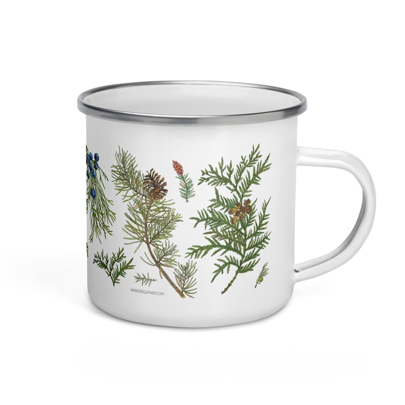May include: White enamel mug featuring a green, brown, and blue botanical illustration of cedar branches, berries, and cones. The mug has a silver rim and a black handle. The text "wandergather.com" is printed on the bottom of the mug.