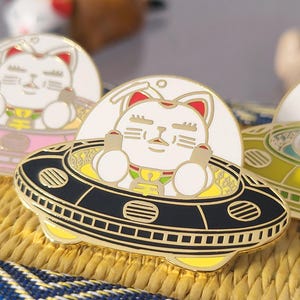 Può includere: Spille smaltate raffiguranti un gatto bianco con orecchie rosse e un collare dorato, seduto in un UFO. Gli UFO sono neri, rosa e gialli con dettagli dorati. Il gatto fa il pollice in su.