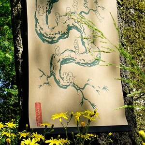 May include: A long, vertical scroll featuring a teal illustration of a winding tree-like form with small figures. The artwork is printed on a tan background and hangs against a tree trunk. The scroll is framed with a black bar at the top.
