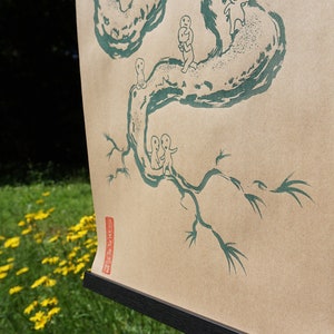 May include: A tan paper print featuring a green illustration of a tree with small figures. The artwork is displayed on a black frame, set against a backdrop of green grass and yellow flowers.