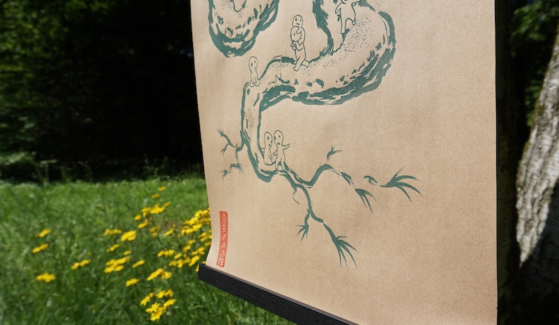 May include: A tan paper print featuring a green illustration of a tree with small figures. The artwork is displayed on a black frame, set against a backdrop of green grass and yellow flowers.