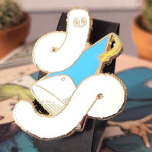 May include: A whimsical enamel pin featuring a white, worm-like creature with a gold outline and a light blue accent. The pin has a gold border and is set against a black background. The design is cartoonish and playful, suitable for accessories.