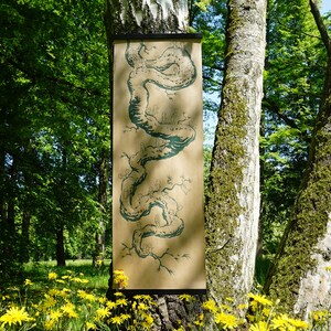 May include: A long, rectangular artwork with a teal illustration of a winding, tree-like form is displayed on a tree trunk. The artwork is on a tan background and is framed by a black bar at the top. The scene is set in a lush green forest.