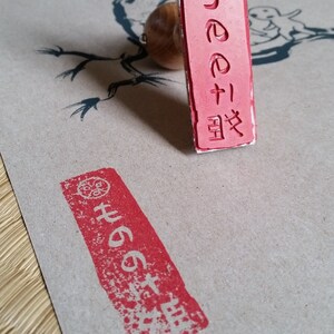 May include: A red Japanese stamp with a wooden handle, next to a stamped impression on tan paper. The stamp features Japanese characters and a circular design. The paper has a dragon illustration.