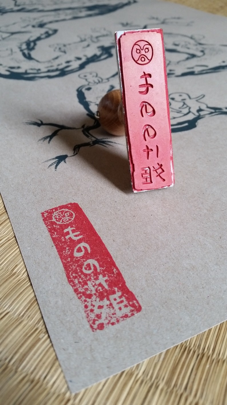 May include: A red Japanese stamp with a wooden handle, next to a stamped impression on tan paper. The stamp features Japanese characters and a circular design. The paper has a dragon illustration.