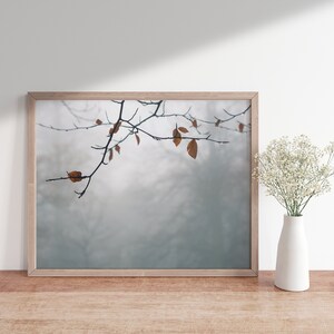 May include: A framed photograph of a branch with brown leaves against a foggy background. The photograph is displayed on a wooden table with a vase of white flowers.
