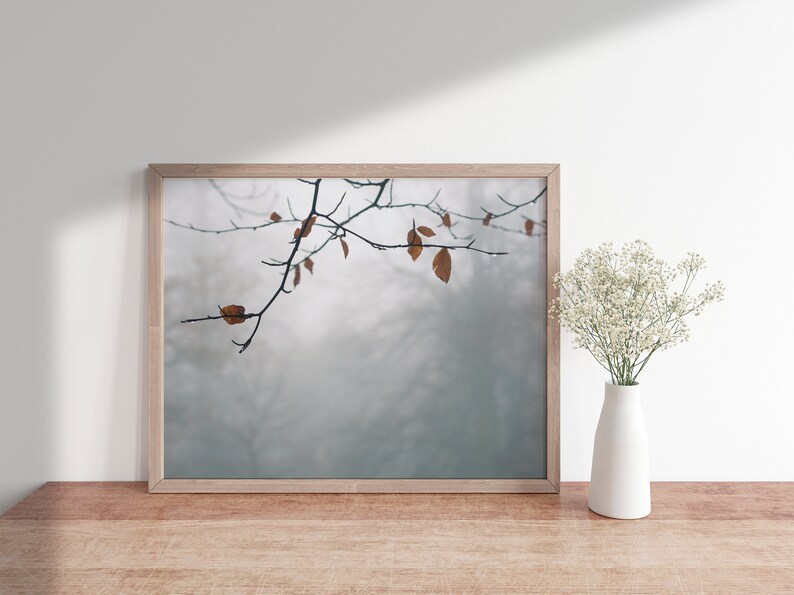 May include: A framed photograph of a branch with brown leaves against a foggy background. The photograph is displayed on a wooden table with a vase of white flowers.