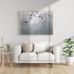 May include: A white sofa with two pillows and a throw blanket sits in a living room with a framed print of a branch with leaves against a foggy background. A small table with a vase of flowers and a potted plant are also in the room.
