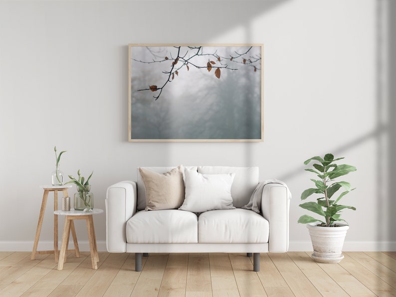 May include: A white sofa with two pillows and a throw blanket sits in a living room with a framed print of a branch with leaves against a foggy background. A small table with a vase of flowers and a potted plant are also in the room.