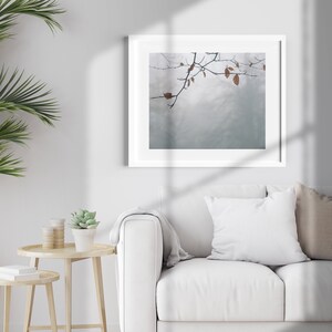 May include: A white framed picture with a muted blue background and a branch with brown leaves hanging from the top. The picture is hanging on a white wall above a white couch with a white pillow and a tan pillow.