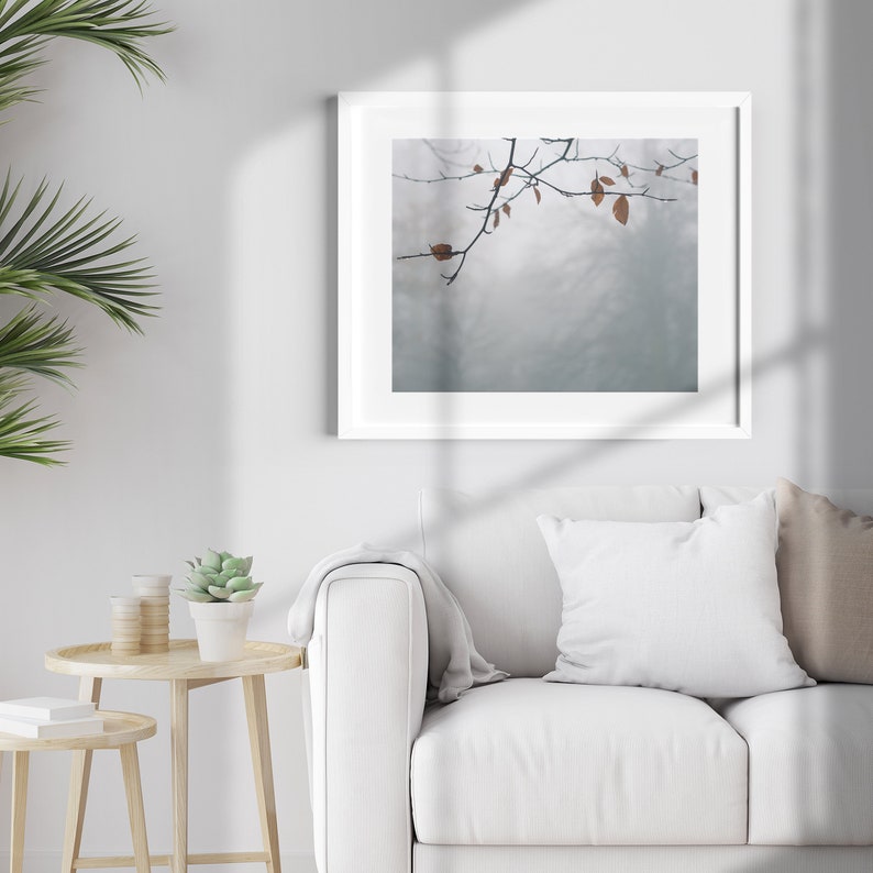 May include: A white framed picture with a muted blue background and a branch with brown leaves hanging from the top. The picture is hanging on a white wall above a white couch with a white pillow and a tan pillow.
