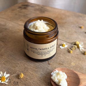May include: A brown glass jar with a white label that reads "nerve/sciatica relief". The jar is filled with a white balm and is sitting on a wooden surface. There are small white flowers scattered around the jar.