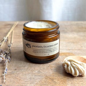 May include: A small amber glass jar with a white label that reads "psoriasis/eczema cream". The jar is filled with a white cream and is sitting on a wooden surface. A small wooden spoon with a dollop of cream is next to the jar.