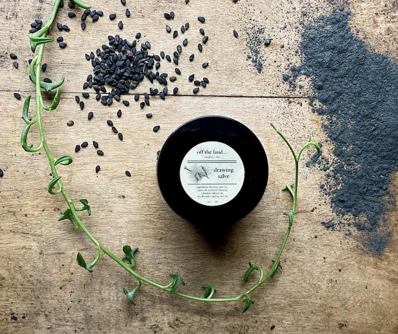 May include: A black jar with a white label reading "drawing salve" sits on a wooden surface, surrounded by black seeds, dark powder, and a green vine. The label features a botanical illustration.