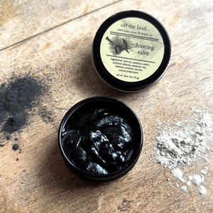 May include: Two small black containers of drawing salve are open on a wooden surface. One container is filled with a dark, thick substance. The other container has a label that reads "off the land... drawing salve." A pile of black powder and white powder are also on the surface.