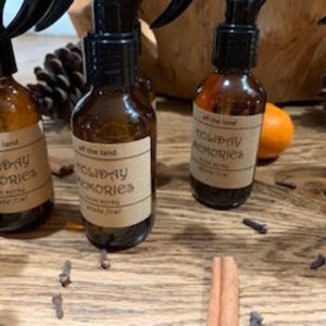 May include: Five amber glass bottles with black spray tops and labels that read "Off the Land Holiday Memories Room Spray". The bottles are arranged in a row on a wooden surface with cinnamon sticks and cloves scattered around them.