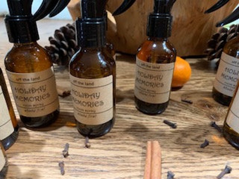May include: Five amber glass bottles with black spray tops and labels that read "Off the Land Holiday Memories Room Spray". The bottles are arranged in a row on a wooden surface with cinnamon sticks and cloves scattered around them.