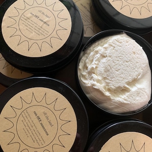 May include: A close-up of three black jars of sun OFF sunscreen with a brown label featuring a sun with rays. The label text reads "off the land" and "sun OFF sunscreen". The ingredients are listed on the label: shea butter, jojoba, beeswax, bazine de oud-carrot seed, red raspberry, hempseed, lemon eucalyptus, lavender, teatree, nitruacile. The jars are filled with a white cream.