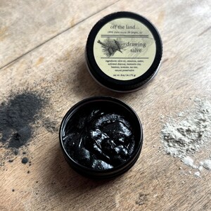 May include: Two small, black metal tins of drawing salve are displayed on a wooden surface. One tin is open, revealing a dark, thick substance. The other tin is closed, with a label that reads "drawing salve" and lists ingredients. Beside the tins are piles of dark and light powders.