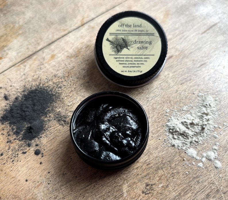 May include: Two small, black metal tins of drawing salve are displayed on a wooden surface. One tin is open, revealing a dark, thick substance. The other tin is closed, with a label that reads "drawing salve" and lists ingredients. Beside the tins are piles of dark and light powders.