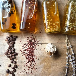 May include: Five glass bottles filled with infused oil, each with a different ingredient: coffee beans, red pepper flakes, garlic, lavender, and vanilla beans. The ingredients are scattered around the bottles on a wooden surface.
