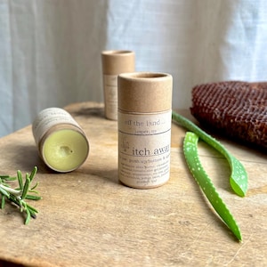 May include: Three cylindrical cardboard tubes of "itch away" balm are displayed on a wooden surface, one open, revealing a yellow balm. The tubes have labels with text. A sprig of rosemary and aloe vera leaves are also present.