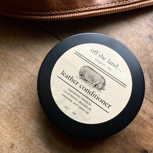 May include: A black jar with a white label that reads "off the land... jasper, ny" and "leather conditioner". The label also lists the ingredients: beeswax, cocoa butter, almond oil, ammonia, and castor oil.