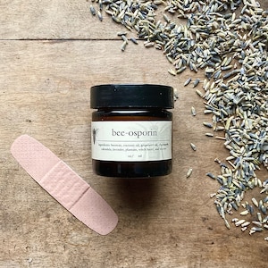 May include: A small glass jar with a brown lid and a white label that reads "bee-osporin". The label lists ingredients including beeswax, coconut oil, grapeseed oil, chamomile, calendula, lavender, plantain, witch hazel, and tea tree.