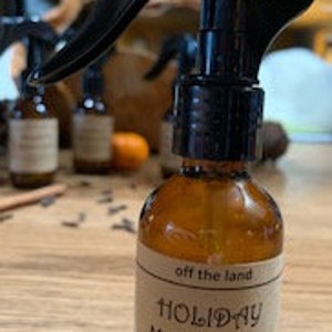 May include: A brown glass bottle with a black spray nozzle filled with room spray. The label reads "off the land HOLIDAY MEMORIES room spray #holidayfirst"