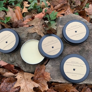 May include: Three small round metal tins with black lids and labels that say "off the land Cast Iron Seasoning Wax" on a bed of brown leaves. The tins are filled with a light yellow wax.