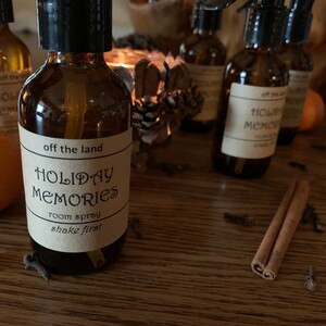 May include: A brown glass bottle with a black spray top and a label that reads "off the land HOLIDAY MEMORIES room spray shake first". The bottle is surrounded by cinnamon sticks, cloves, and oranges.