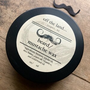 May include: A black container of beard and mustache wax with a cream-colored label. The label features a stylized mustache graphic and the text "off the land..." and "beard/ mustache wax". A small, black mustache cutout is next to the container.