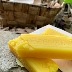 May include: Close-up of three yellow beeswax bars with the word "Beeswax" embossed on them. The bars have a floral design and are resting on a stone surface. A woven basket is in the background.