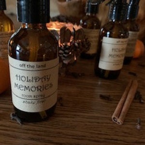 May include: A brown glass bottle with a black spray nozzle and a white label that reads "off the land HOLIDAY MEMORIES ROOM SPRAY shake well". The bottle is sitting on a wooden surface with a few oranges, cinnamon sticks, and cloves.